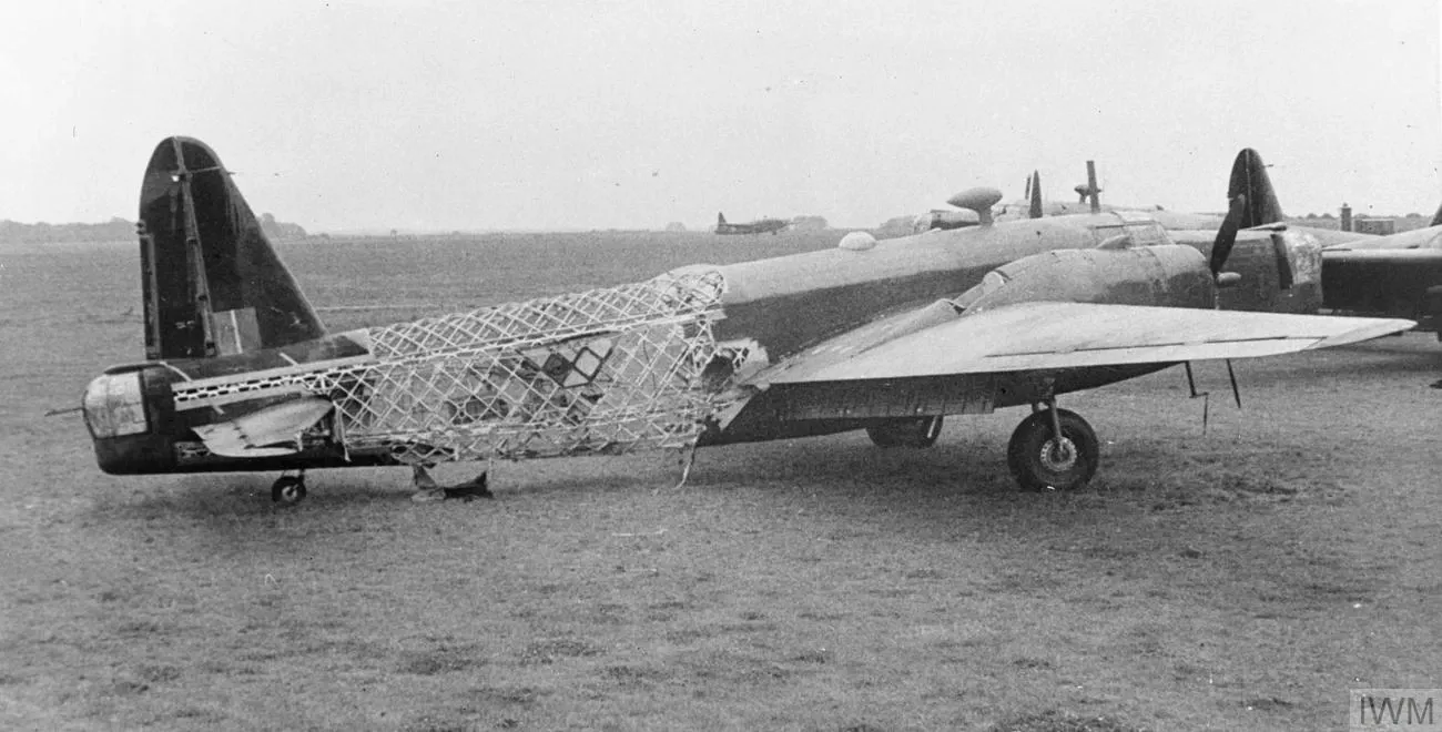 A Vickers Wellington bomber standing on grass at an airfield, the fabric torn away from its rear fuselage to expose the bare lattice of its geodetic frame. Other aircraft stand behind it.